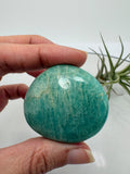 palmstone de amazonita natural pulida con inclusiones de cuarzo ahumado y mica biotita, de Madagascar.

&nbsp;Una piedra perfecta para llevar en la mano, meditar o utilizar como apoyo energético diario. La amazonita destaca por sus tonos verde azulados con vetas naturales más oscuras, que le aportan profundidad y un aspecto único.