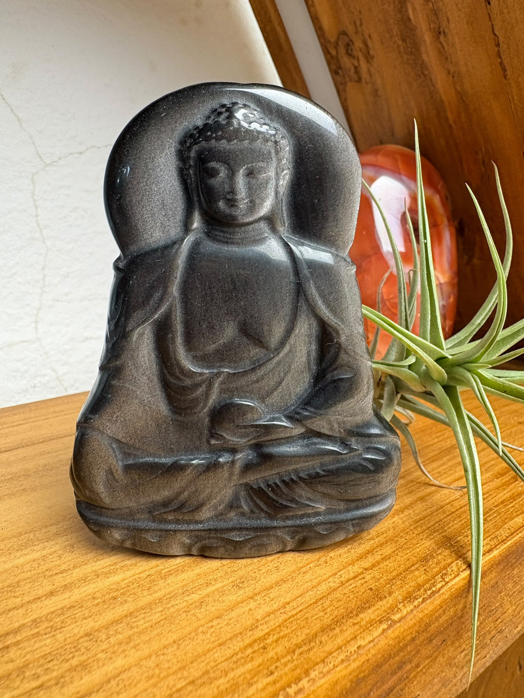 Buda de obsidiana plateada tallado a mano destaca por sus preciosos reflejos plateados sobre un fondo oscuro. La luz realza los pliegues de su vestimenta, su rostro sereno y su postura de meditación, mostrando la belleza de la talla.