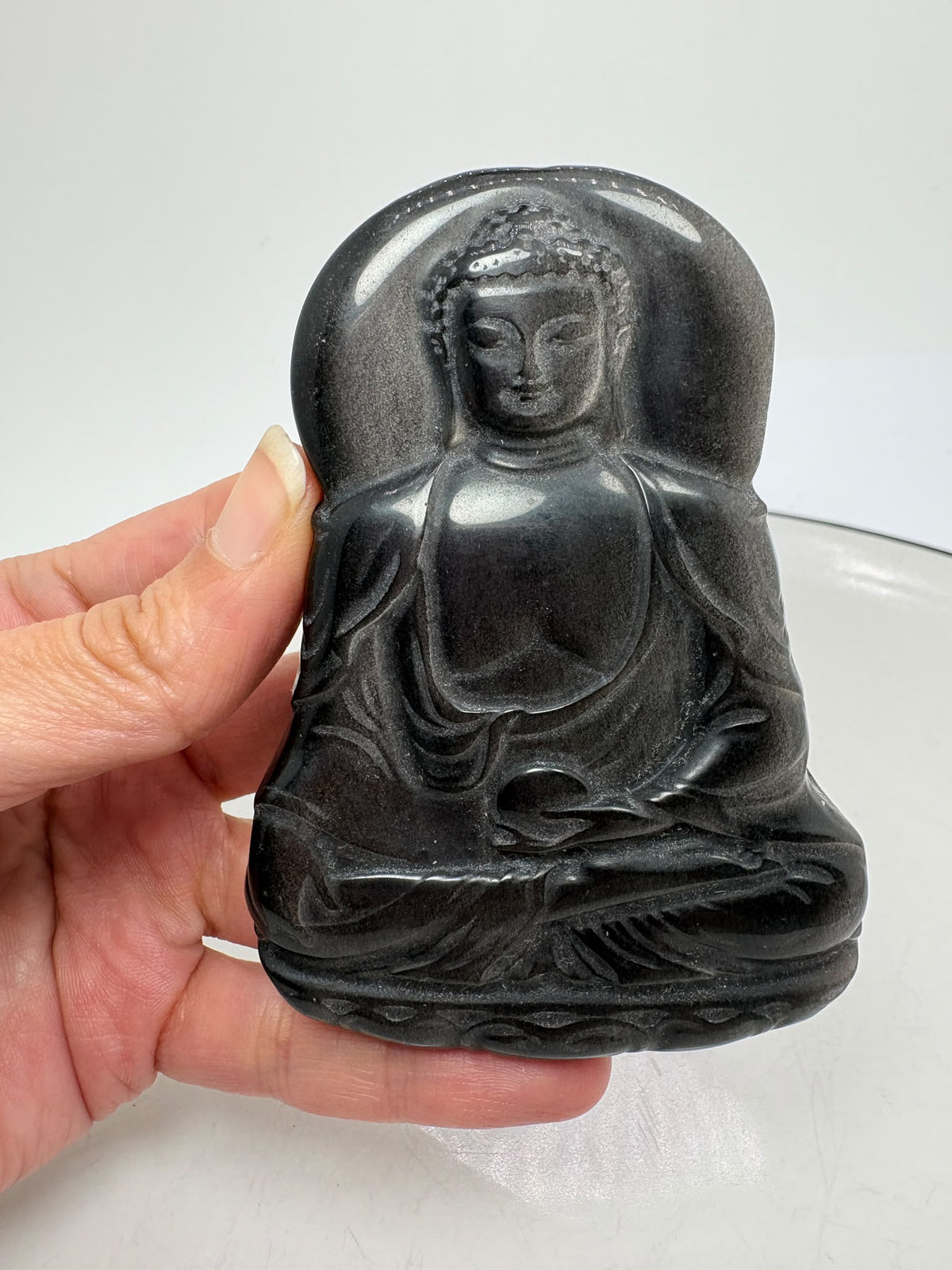 Buda de obsidiana plateada tallado a mano destaca por sus preciosos reflejos plateados sobre un fondo oscuro. La luz realza los pliegues de su vestimenta, su rostro sereno y su postura de meditación, mostrando la belleza de la talla.