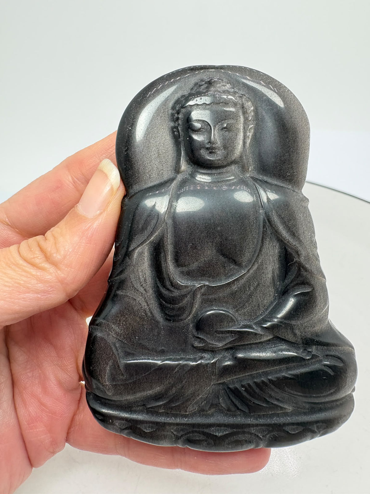 Buda de obsidiana plateada tallado a mano destaca por sus preciosos reflejos plateados sobre un fondo oscuro. La luz realza los pliegues de su vestimenta, su rostro sereno y su postura de meditación, mostrando la belleza de la talla.