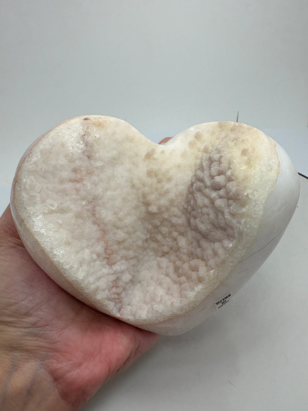 Corazón grande de ágata con una bonita geoda de calcita con micro cristalitos de cuarzo que hace que brillen, en tonos blancos y crema y rosado clarito.

Su cavidad deja a la vista los cristales, con pequeños brillos que se aprecian al moverlo bajo la luz.

Los bordes y la parte trasera están pulidos, mostrando zonas translúcidas y dibujos en tonos tierra. El contraste con la textura de la geoda hace que sea una pieza bonita por ambas caras, ideal para decorar un rincón especial o ampliar tu colección.
