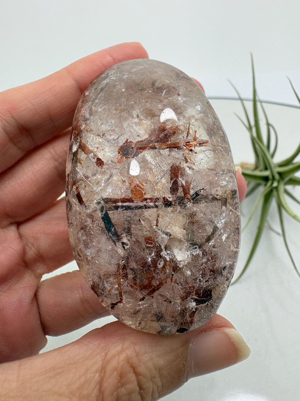Un pequeño paisaje mineral en la palma de tu mano. Este palmstone de cuarzo con anfíbol destaca por sus inclusiones en tonos verdes oscuros, marrones y rojizos, que crean formas y contrastes únicos en su interior.

La transparencia del cuarzo permite descubrir distintos detalles al observarlo desde diferentes ángulos. Su acabado semipulido combina zonas suaves y brillantes con una textura más natural.