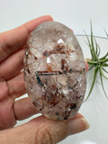 Un pequeño paisaje mineral en la palma de tu mano. Este palmstone de cuarzo con anfíbol destaca por sus inclusiones en tonos verdes oscuros, marrones y rojizos, que crean formas y contrastes únicos en su interior.

La transparencia del cuarzo permite descubrir distintos detalles al observarlo desde diferentes ángulos. Su acabado semipulido combina zonas suaves y brillantes con una textura más natural.