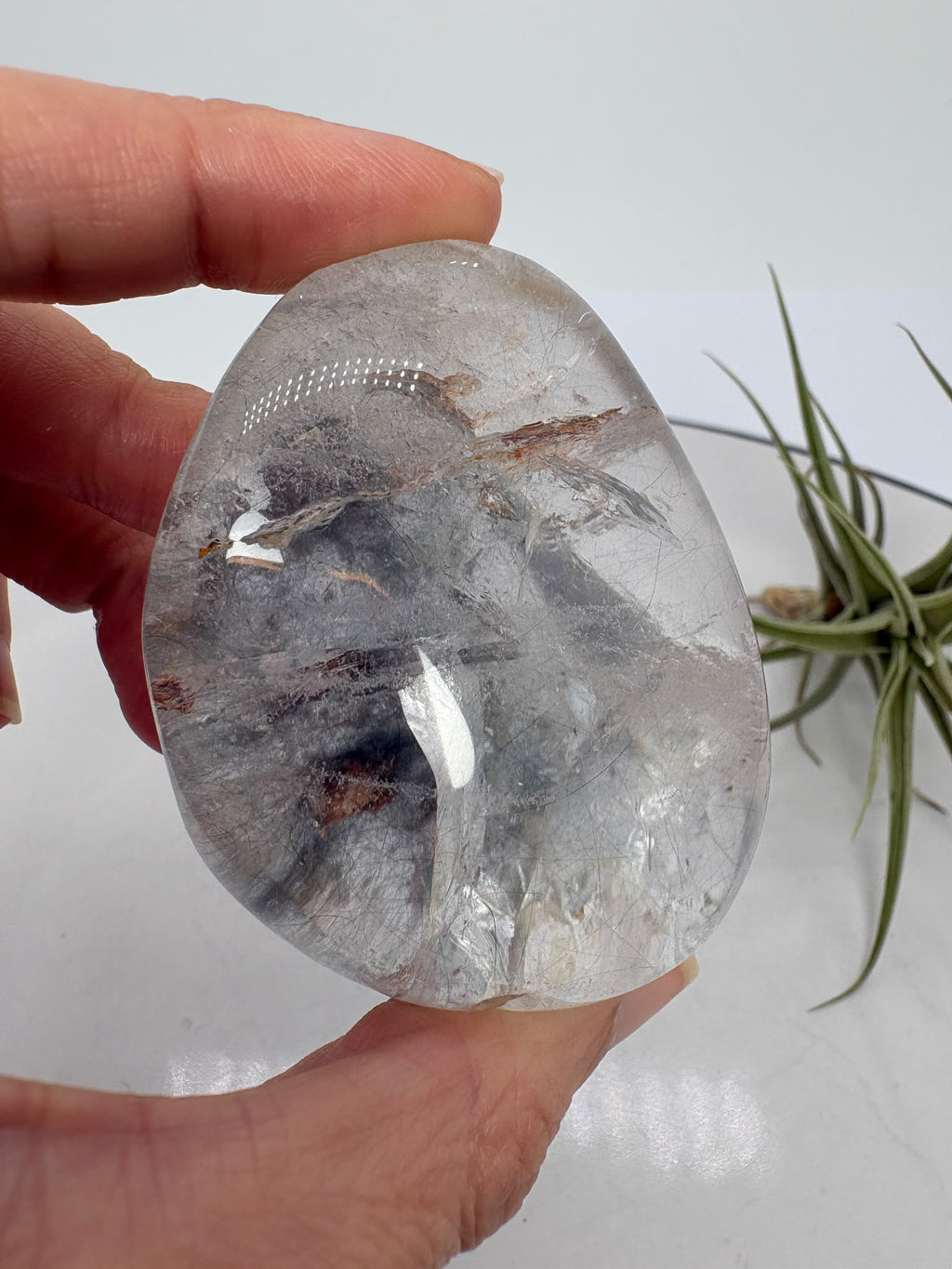 Un pequeño universo natural en el interior del cuarzo. Este palmstone contiene finas agujas de rutilo e inclusiones de aspecto plateado, que crean un paisaje lleno de detalles entre transparencias y delicados matices cálidos.
