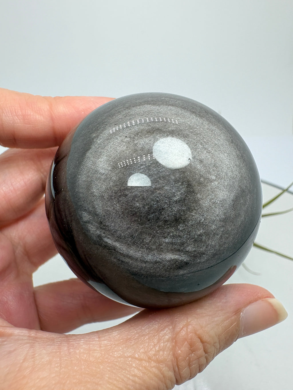 Esfera de obsidiana plateada con un bonito brillo que se aprecia al moverla bajo la luz. Sobre su color negro destacan reflejos plateados que cambian según el ángulo desde el que la mires, dejando ver sus dibujos naturales.
