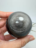Esfera de obsidiana plateada con un bonito brillo que se aprecia al moverla bajo la luz. Sobre su color negro destacan reflejos plateados que cambian según el ángulo desde el que la mires, dejando ver sus dibujos naturales.
