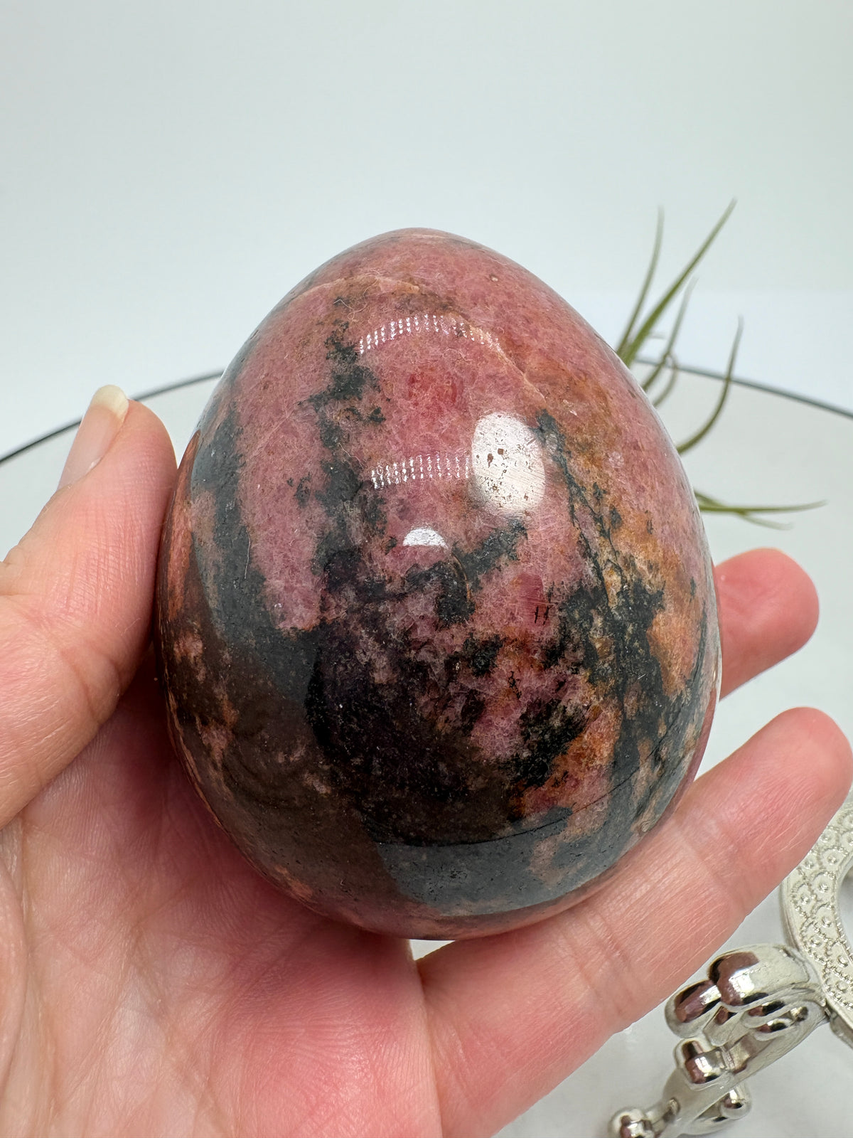 Un precioso huevo de rodonita que combina tonos rosa intenso con zonas negras y matices terrosos. Sus dibujos cambian en cada cara, mostrando una parte más rosada y otra más oscura, con un contraste que le da mucha personalidad. El acabado pulido realza sus colores y su brillo natural.