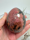 Un precioso huevo de rodonita que combina tonos rosa intenso con zonas negras y matices terrosos. Sus dibujos cambian en cada cara, mostrando una parte más rosada y otra más oscura, con un contraste que le da mucha personalidad. El acabado pulido realza sus colores y su brillo natural.