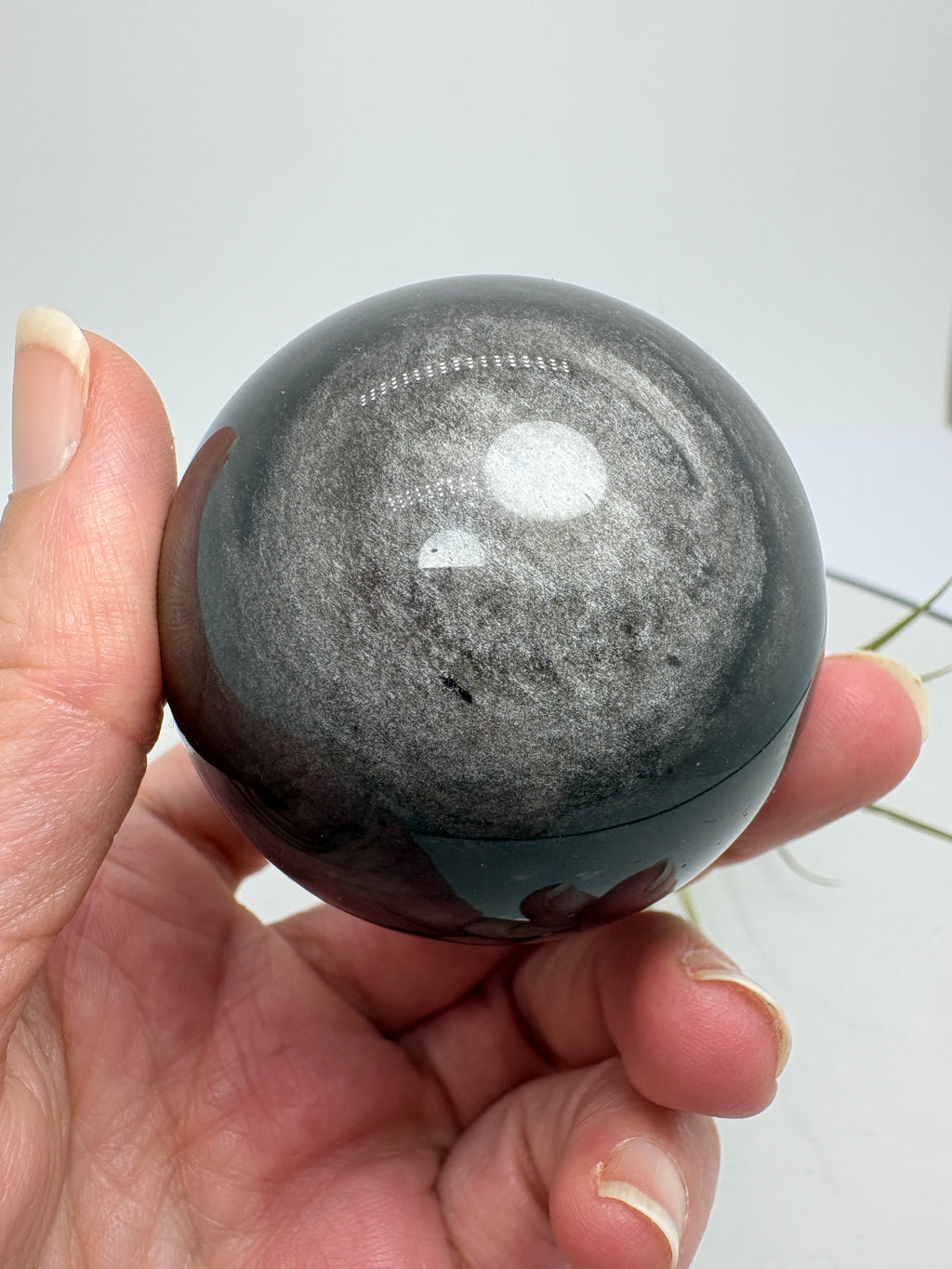 Esfera de obsidiana plateada con un bonito brillo que se aprecia al moverla bajo la luz. Sobre su color negro destacan reflejos plateados que cambian según el ángulo desde el que la mires, dejando ver sus dibujos naturales.