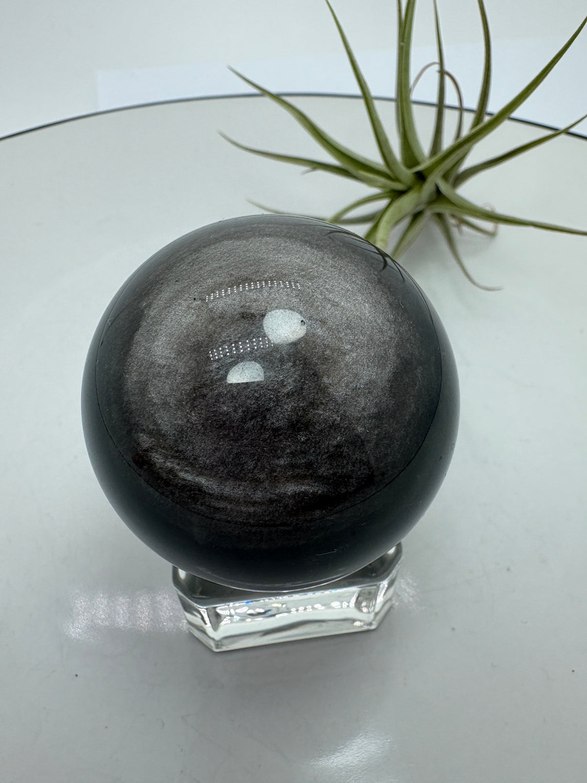 Esfera de obsidiana plateada con un bonito brillo que se aprecia al moverla bajo la luz. Sobre su color negro destacan reflejos plateados que cambian según el ángulo desde el que la mires, dejando ver sus dibujos naturales.