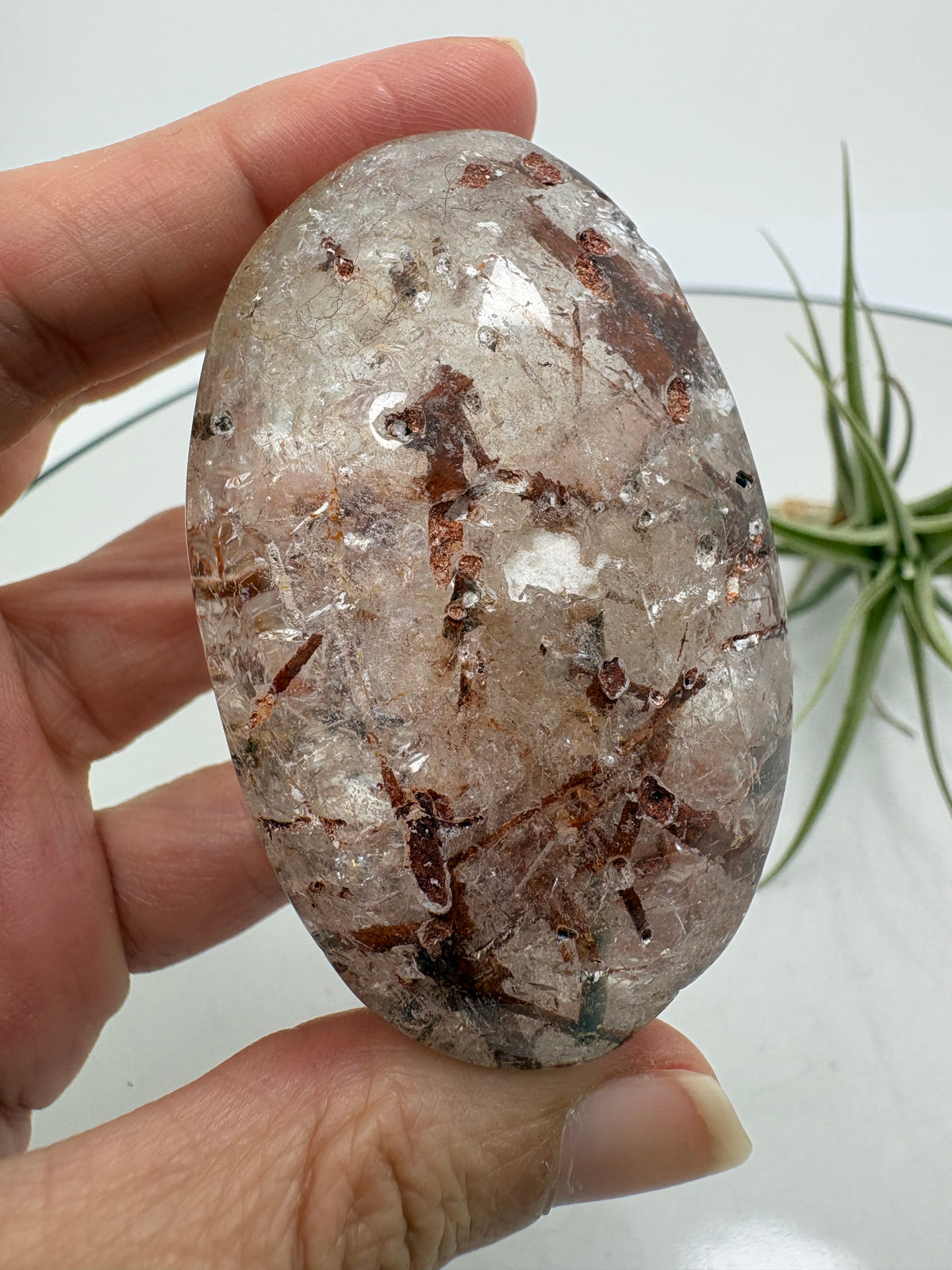 Un pequeño paisaje mineral en la palma de tu mano. Este palmstone de cuarzo con anfíbol destaca por sus inclusiones en tonos verdes oscuros, marrones y rojizos, que crean formas y contrastes únicos en su interior.

La transparencia del cuarzo permite descubrir distintos detalles al observarlo desde diferentes ángulos. Su acabado semipulido combina zonas suaves y brillantes con una textura más natural.
