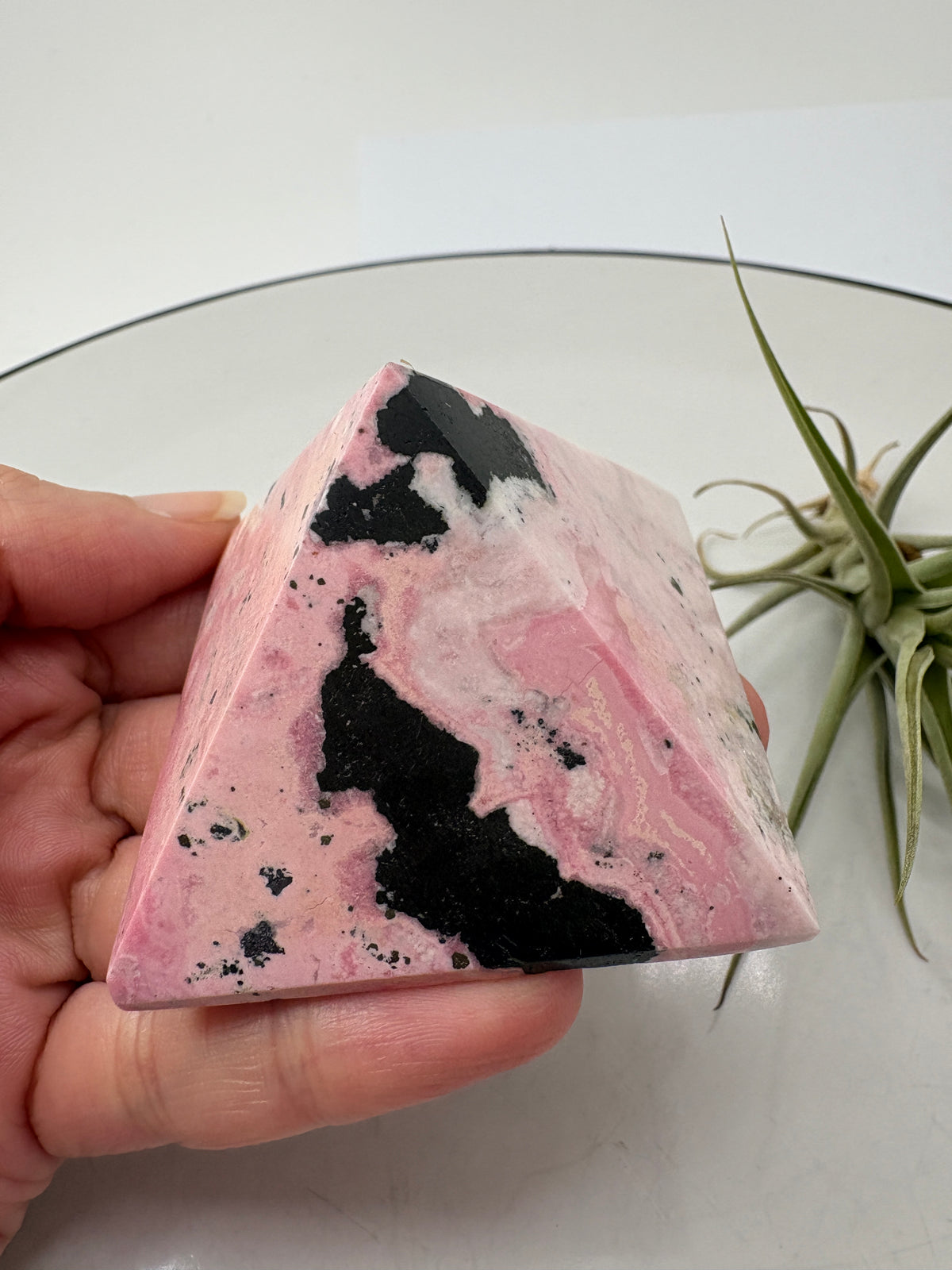 Pirámide de rodonita de Perú con una bonita combinación de tonos rosas, zonas claras y manchas negras que destacan sobre su superficie pulida. Sus dibujos naturales hacen de ella una pieza única, ideal para decorar un rincón especial o ampliar tu colección de minerales.