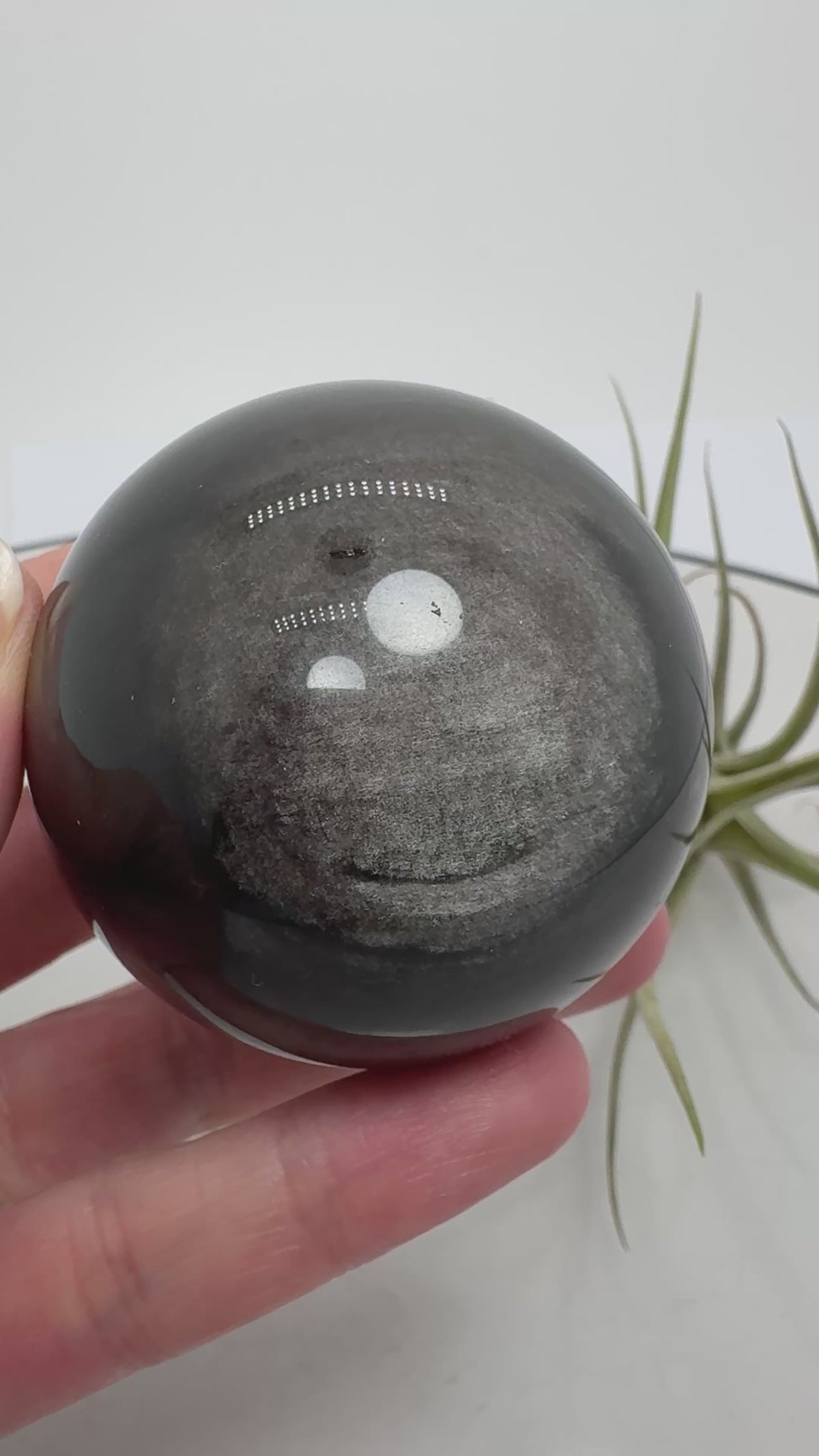 Esfera de obsidiana plateada con un bonito brillo que se aprecia al moverla bajo la luz. Sobre su color negro destacan reflejos plateados que cambian según el ángulo desde el que la mires, dejando ver sus dibujos naturales.