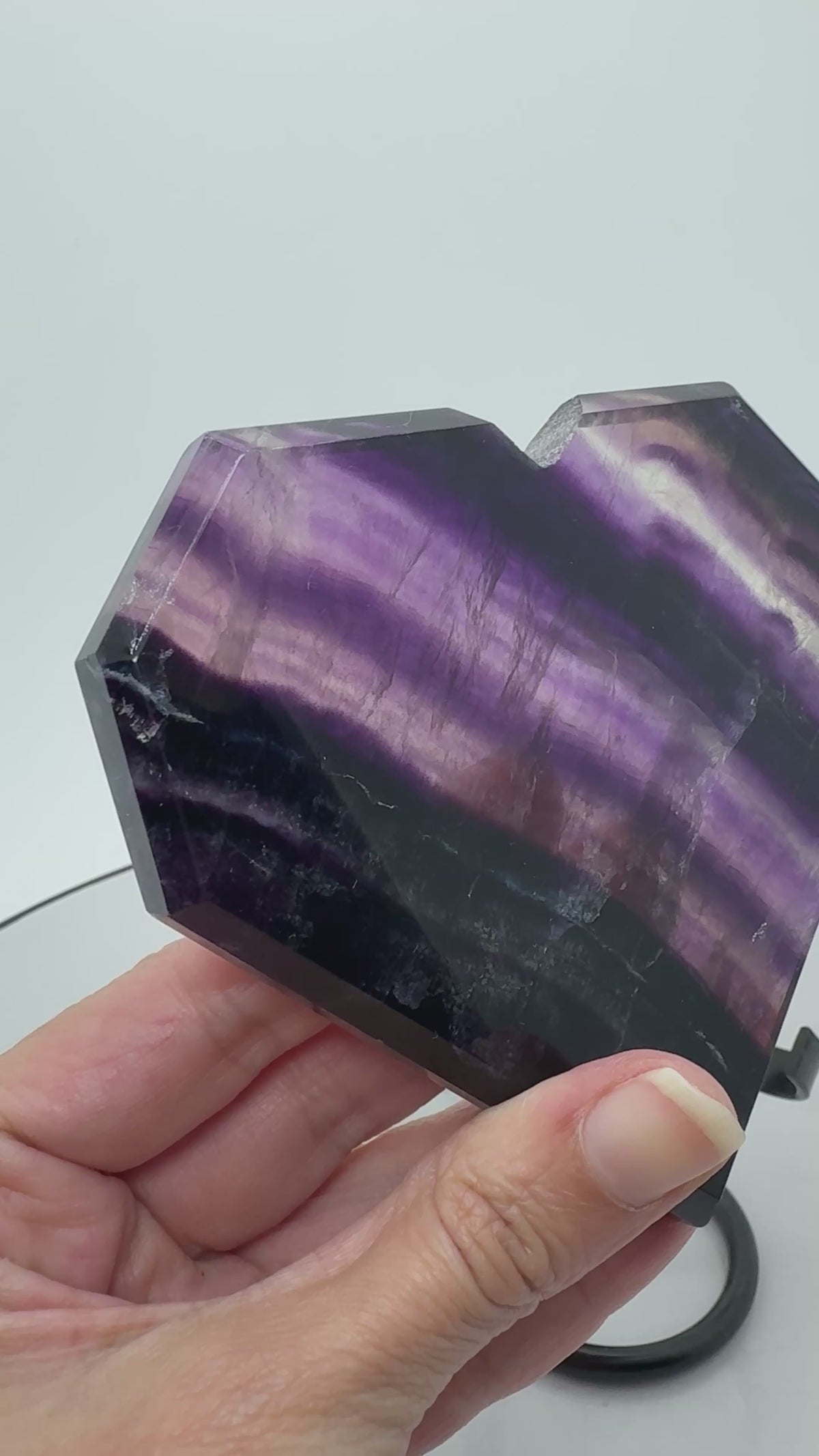Un corazón de fluorita arcoíris con bonitas bandas violetas, zonas translúcidas y un ligero toque azul que se aprecia entre sus vetas. Sus tonos se realzan al pasar la luz a través de la pieza, dejando ver los dibujos de su interior.
