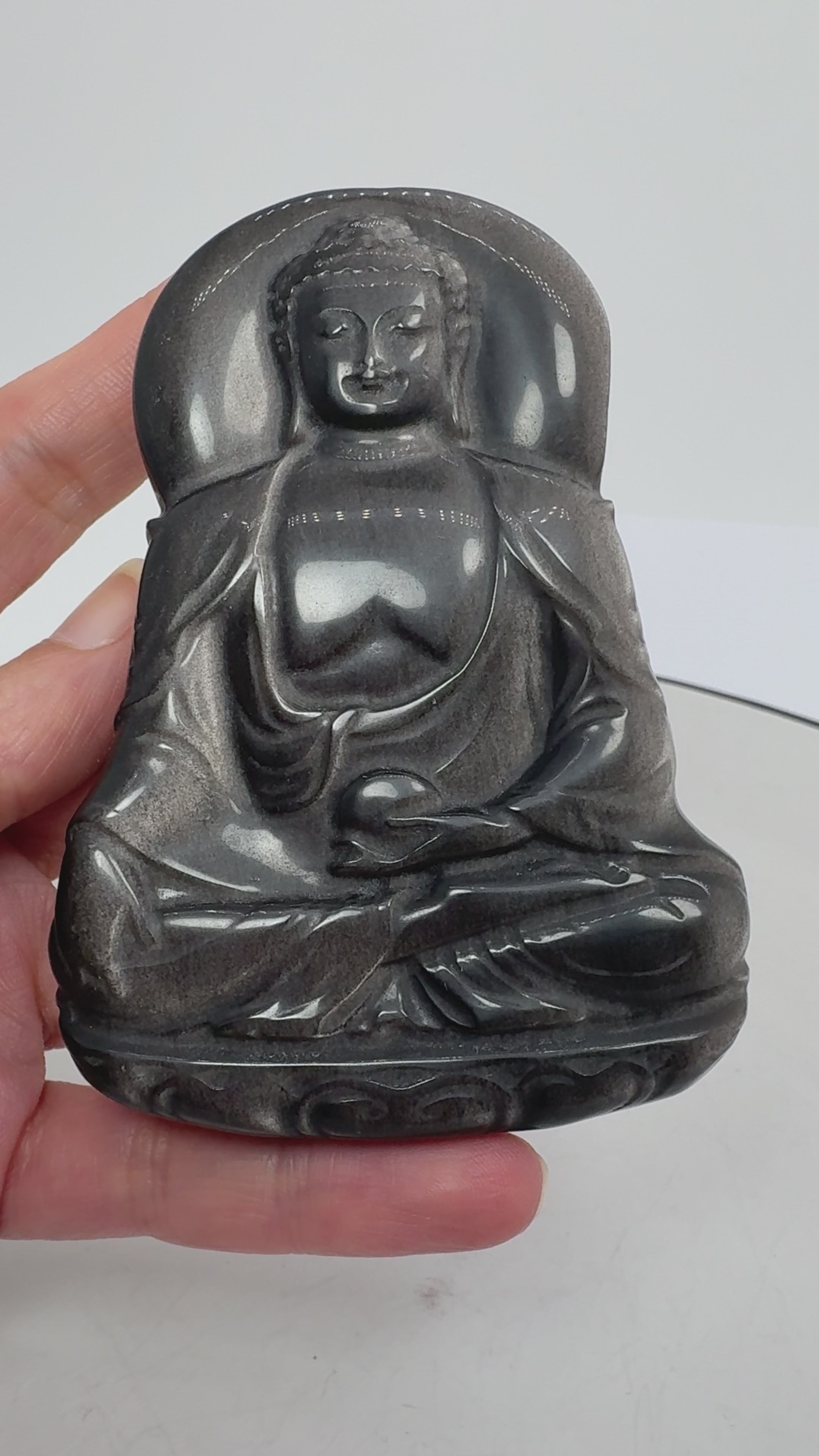 Buda de obsidiana plateada tallado a mano destaca por sus preciosos reflejos plateados sobre un fondo oscuro. La luz realza los pliegues de su vestimenta, su rostro sereno y su postura de meditación, mostrando la belleza de la talla.